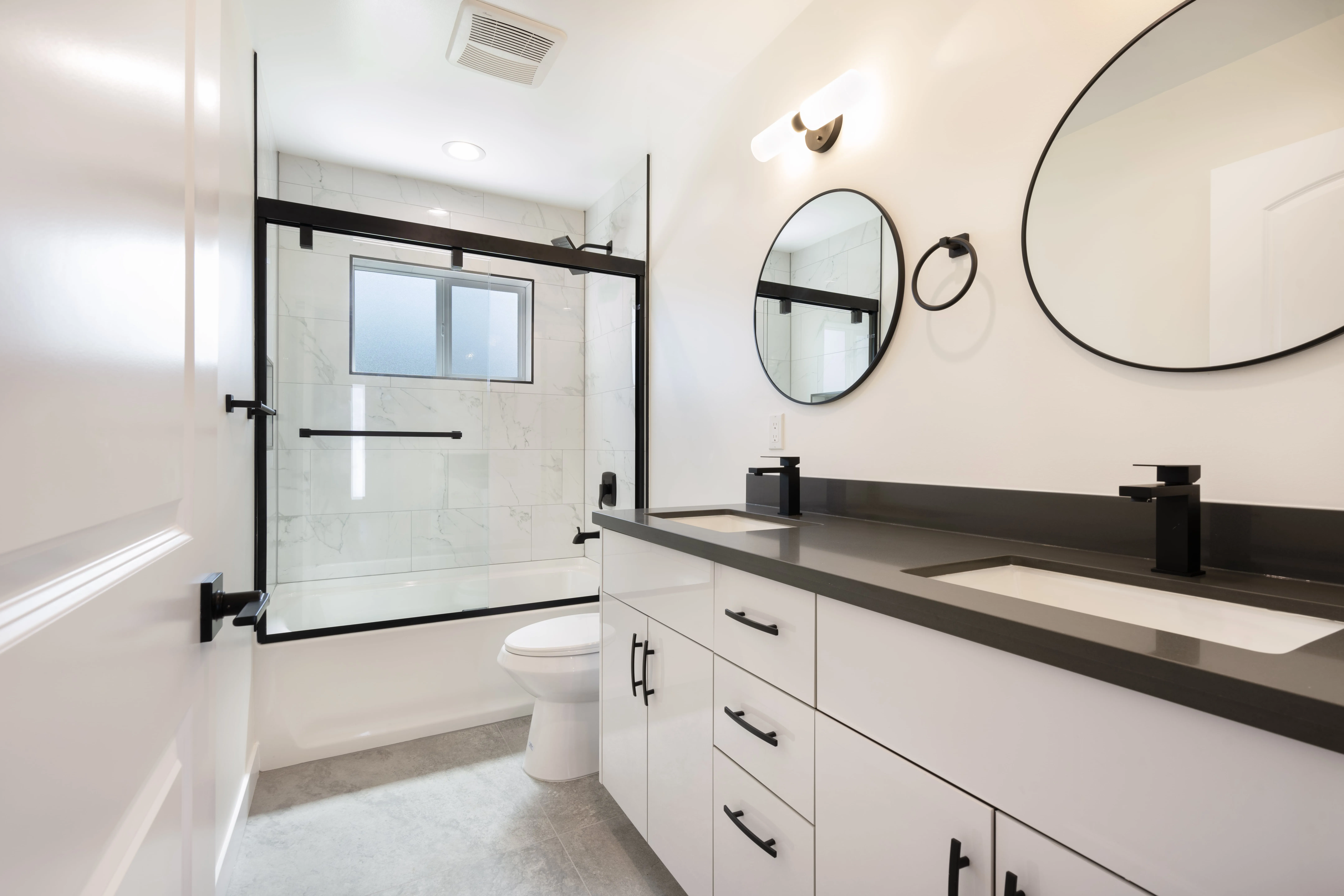 Luxury ADU bathroom with dual vanity, round mirrors, marble tile shower, and modern black fixtures