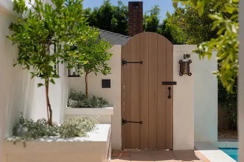 Arched wooden garden gate framed by white stucco walls, lanterns, and greenery